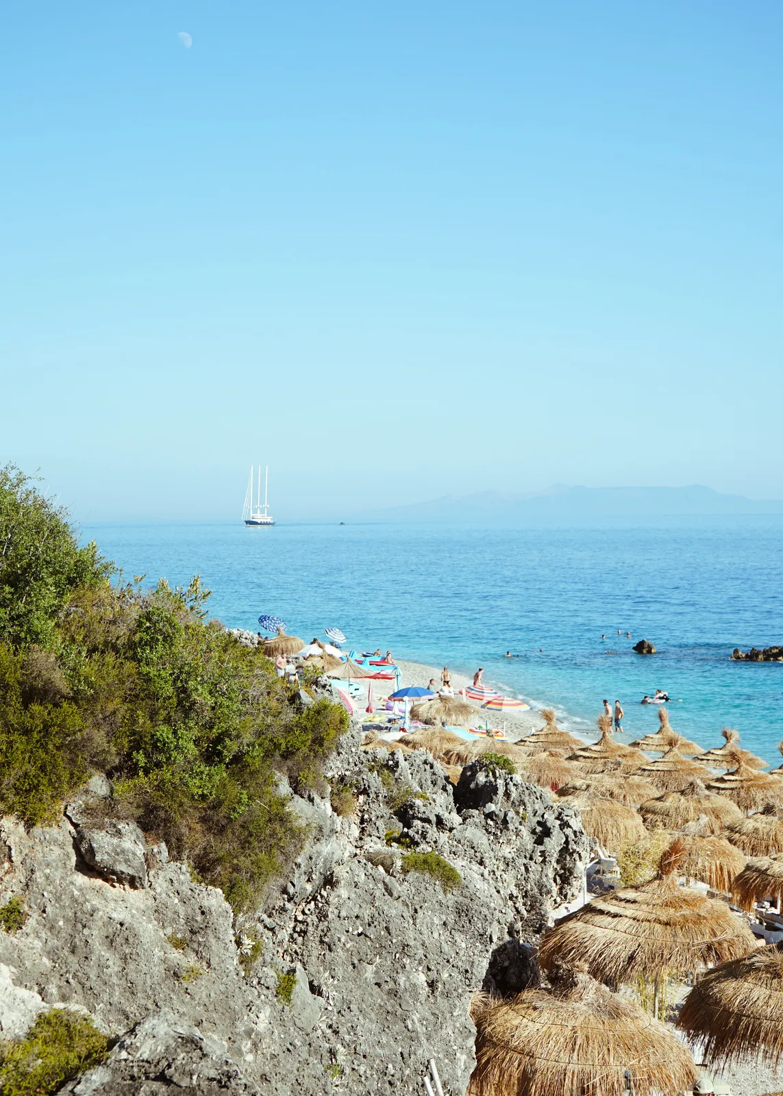 Dhërmi Albania — rows of thatched straw umbrellas along a pebble beach with a tall sailboat anchored on the calm blue Ionian and mountains beyond, Albanian Riviera