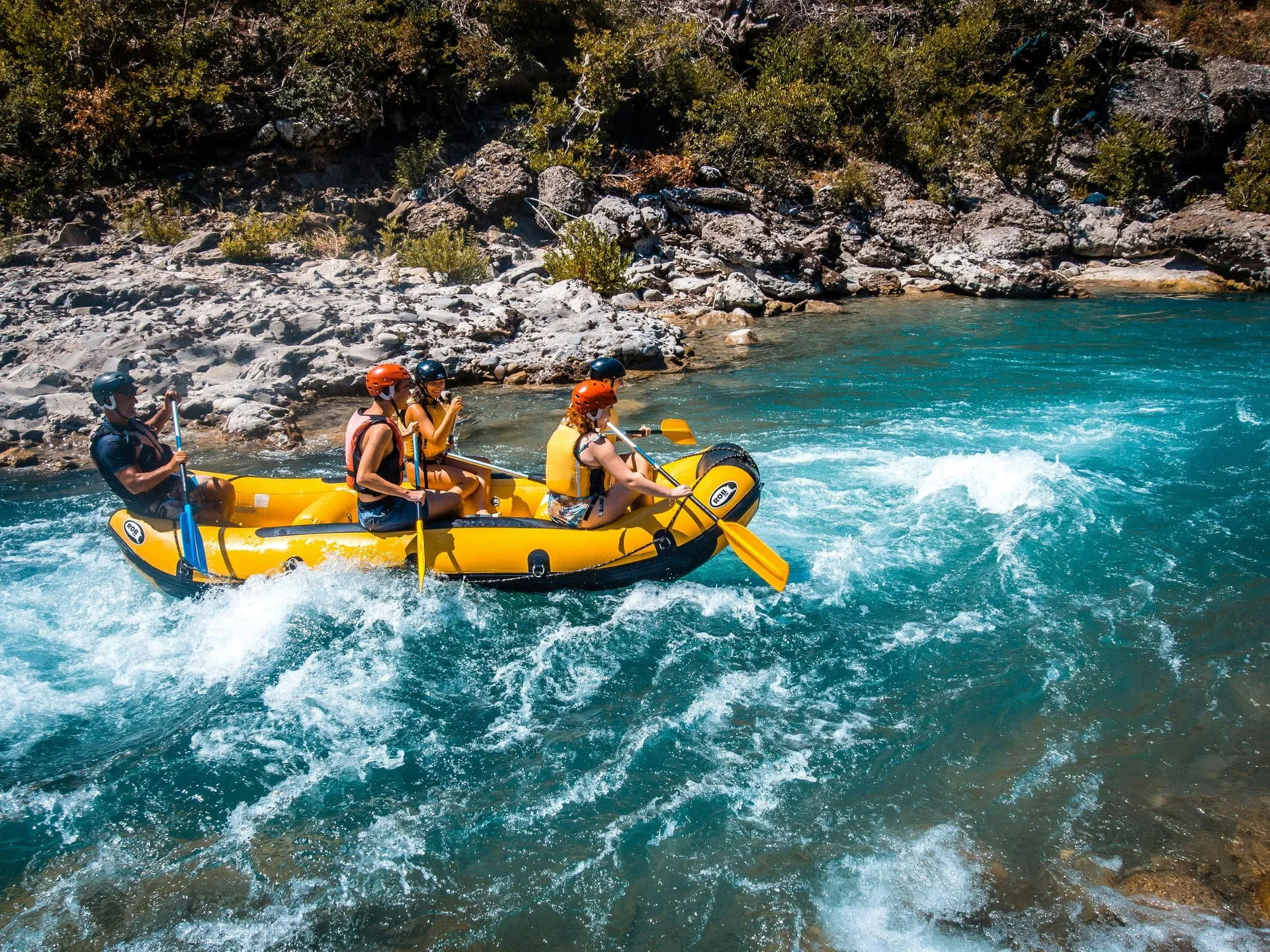 Yellow inflatable raft with four people in helmets paddling through white-water rapids on the vivid turquoise Vjosa River, rocky banks and green hills behind, Përmet Albania
