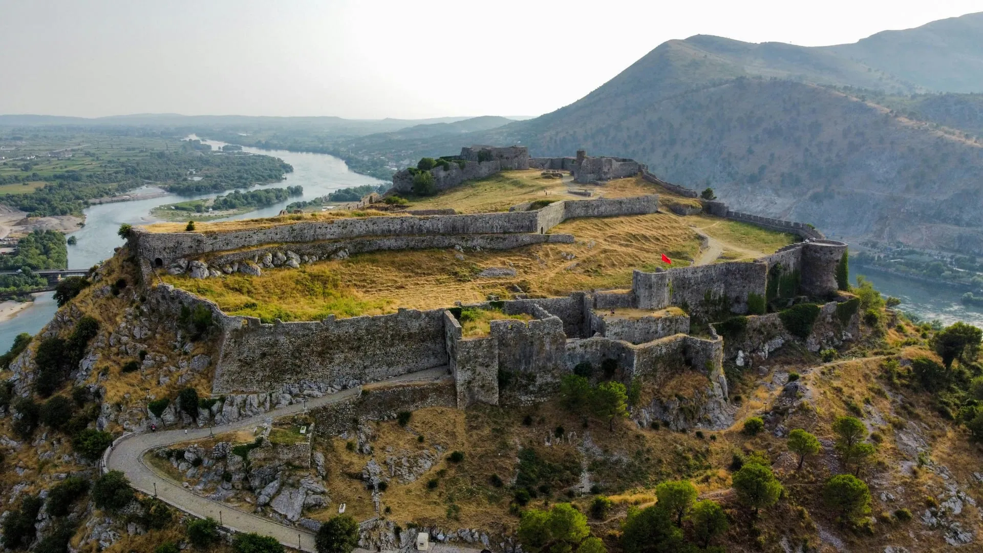 Aerial view of Rozafa Castle above Shkodër, its long stone ramparts enclosing grassy courtyards on a hilltop, with rivers winding through the green plain below and mountains behind, Albania